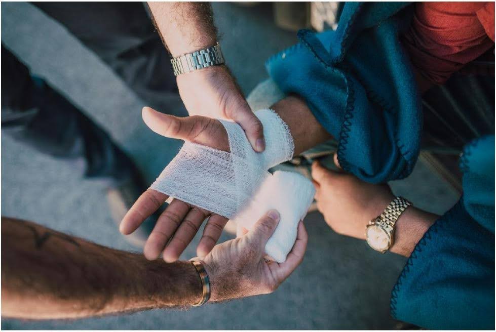 High-angle shot of multiple people's hands working together to wrap a white gauze bandage around another person's wrist and palm.