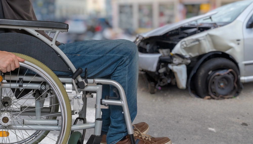Disabled man in a wheelchair after a car accident with a crashed vehicle in the background.