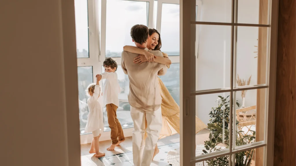 An adult couple and two young children dance joyfully in a sunlit, modern living room with large windows.