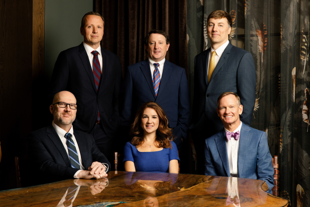 Group portrait of The Clardy Law Firm team, —posed around a polished wood table against a dark, patterned backdrop.