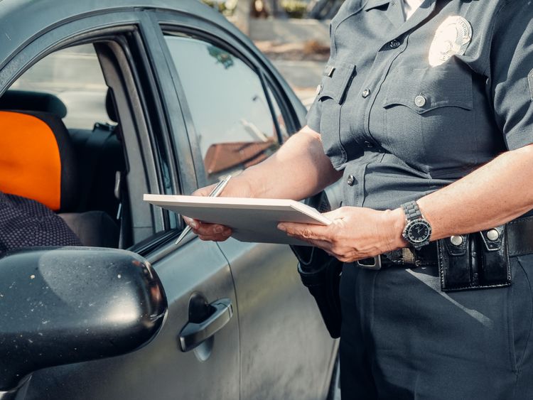A police officer standing by a car window while writing on a clipboard during a traffic stop.